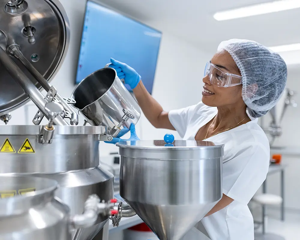 Woman with safety goggles and hair net working in chemical facility adding liquid product to large mixer.