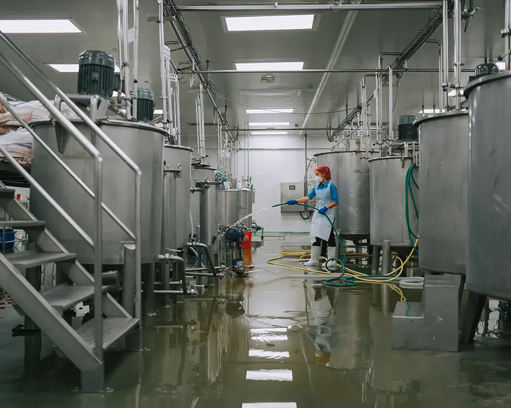Wide shot of a row of elevated industrial containers; a person in safety gear is shown washing the equipment with a stream of liquid coming out of a hose.