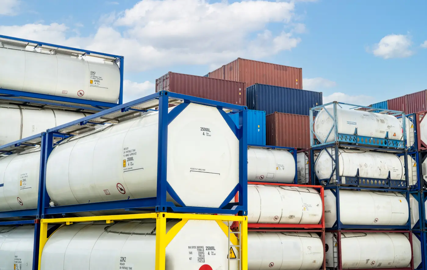 Stacks of white cylindrical tank containers and rectangular cargo containers at a shipping yard under a blue sky with scattered clouds.